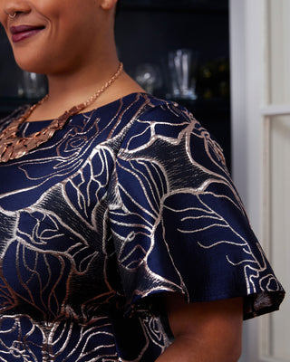 Woman wearing a dark blue dress with gold floral patterns indoors. This dress is the Elmley Sheath Dress sewing pattern from "Sewing the Curve" by Jenny Rushmore.