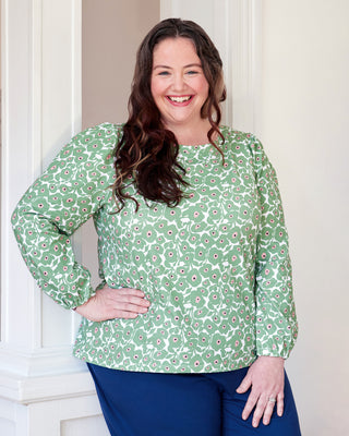Woman wearing a green floral blouse and blue pants standing against a white wall. This top is the Eustis T-Shirt sewing pattern from "Sewing the Curve" by Jenny Rushmore.