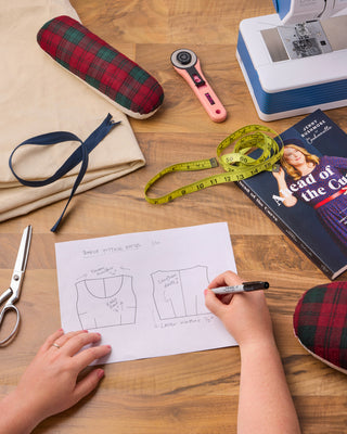 Person drawing a sewing pattern on paper with sewing tools on a wooden table.