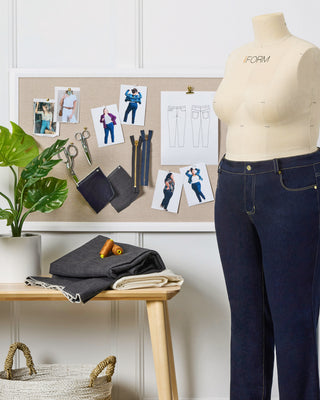 Mannequin wearing dark blue jeans next to a cork board with fashion sketches and a plant.