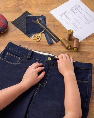 Person working on a pair of blue jeans with sewing tools and materials on a wooden table.