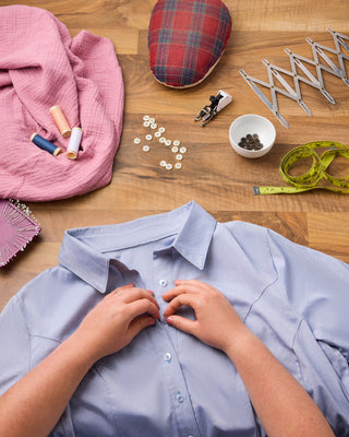 Person sewing a button onto a shirt with sewing supplies on a wooden table