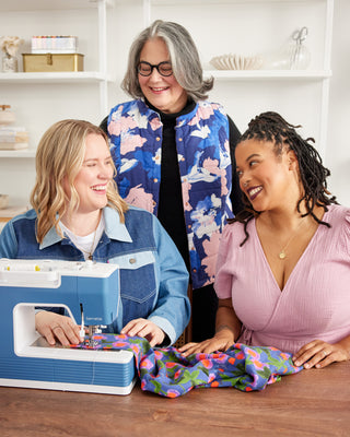 Three women working together at a sewing machine with colorful fabric on a wooden table.