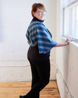 Person wearing a blue and gray plaid shawl in an indoor setting. The topper is the Caroll Shrug sewing pattern from Cashmerette.