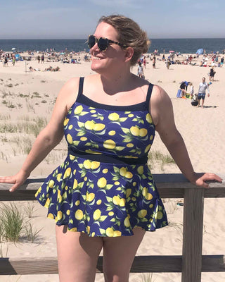 A woman standing on a beach boardwalk wearing a blue and yellow tankini swimsuit with a circular peplum skirt. This swimsuit is the Ipswich Swimsuit sewing pattern from Cashmerette.