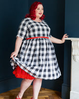 Woman wearing a black and white checkered dress with a red skirt and red shoes, standing against a dark blue wall.  This dress is the Honeybourne Fit and Flare Dress sewing pattern from "Ahead of the Curve" by Jenny Rushmore.