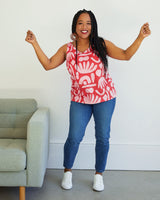 Woman wearing a red patterned sleeveless top and blue jeans standing in a room with a gray couch. This top is the Kersoe Top sewing pattern from "Ahead of the Curve" by Jenny Rushmore.