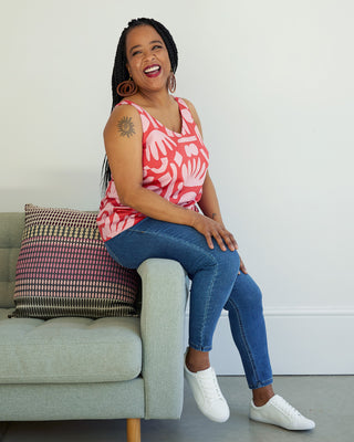 Woman sitting on a couch wearing a red floral tank top and blue jeans, with a neutral background. This top is the Kersoe Top sewing pattern from "Ahead of the Curve" by Jenny Rushmore.