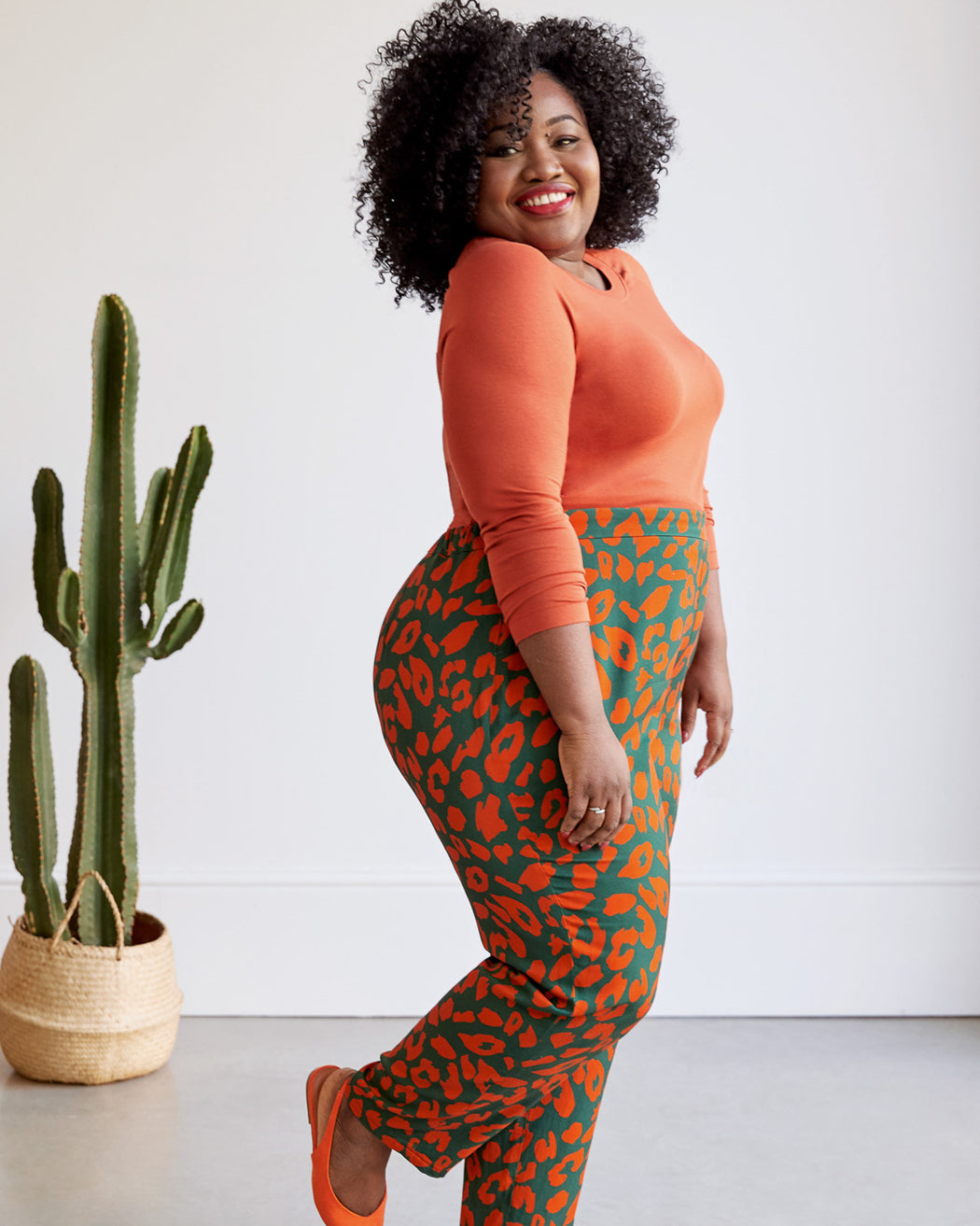Woman wearing an orange top and green leopard print pants standing next to a cactus plant. These pants are made with the Magna Pants sewing pattern from "Ahead of the Curve" by Jenny Rushmore.