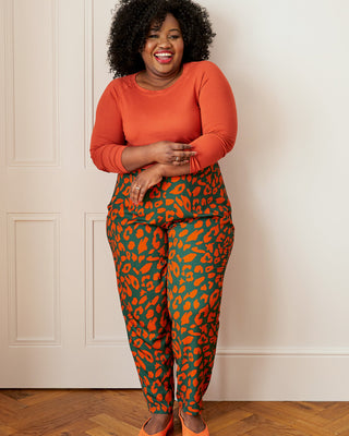 Woman wearing an orange top and green leopard print pants standing against a white wall. These pants are made with the Magna Pants sewing pattern from "Ahead of the Curve" by Jenny Rushmore.