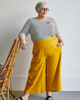 Woman wearing a striped shirt and yellow pants standing next to a wicker chair. This top is the Stanway Raglan T-Shirt sewing pattern from "Ahead of the Curve" by Jenny Rushmore.