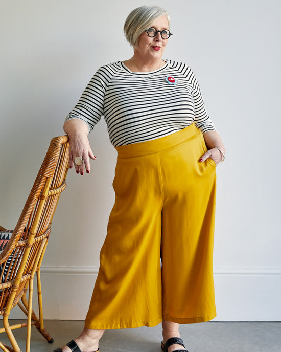 Woman wearing a striped shirt and yellow pants standing next to a wicker chair. This top is the Stanway Raglan T-Shirt sewing pattern from "Ahead of the Curve" by Jenny Rushmore.