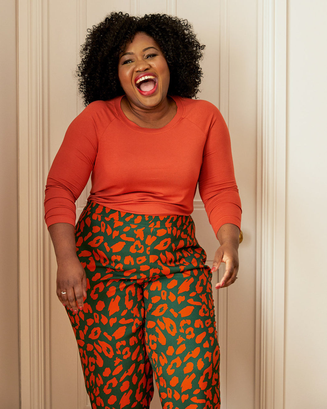 Woman wearing an orange long-sleeve top and green leggings with red leopard print on a white background. This top is the Stanway Raglan T-Shirt sewing pattern from "Ahead of the Curve" by Jenny Rushmore.