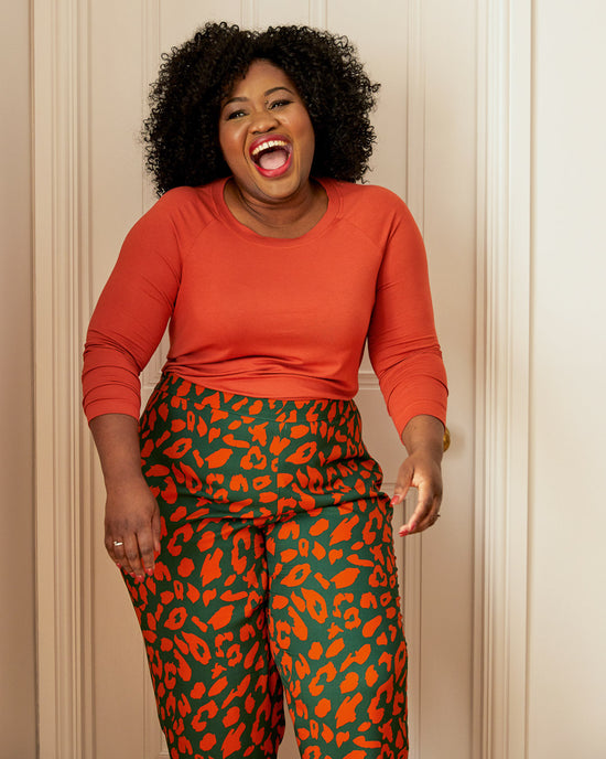 Woman wearing an orange long-sleeve top and green leggings with red leopard print on a white background. This top is the Stanway Raglan T-Shirt sewing pattern from "Ahead of the Curve" by Jenny Rushmore.