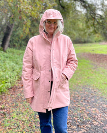 Person wearing a pink raincoat walking on a path with trees in the background. This is the Marston Raincoat sewing pattern.