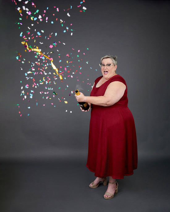 Woman in a red dress holding a bottle with confetti falling around her on a dark background. This dress is the Upton Dress & Skirt sewing pattern from Cashmerette.