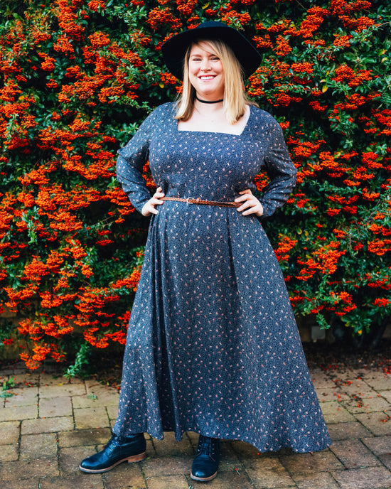 Woman in a blue dress standing in front of a hedge with red flowers. This dress is the Upton Dress & Skirt sewing pattern from Cashmerette.