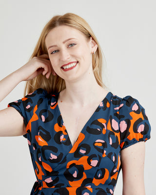 Woman wearing a blue dress with orange and pink abstract pattern on a plain background. This dress is the Roseclair Wrap Dress sewing pattern from Cashmerette.