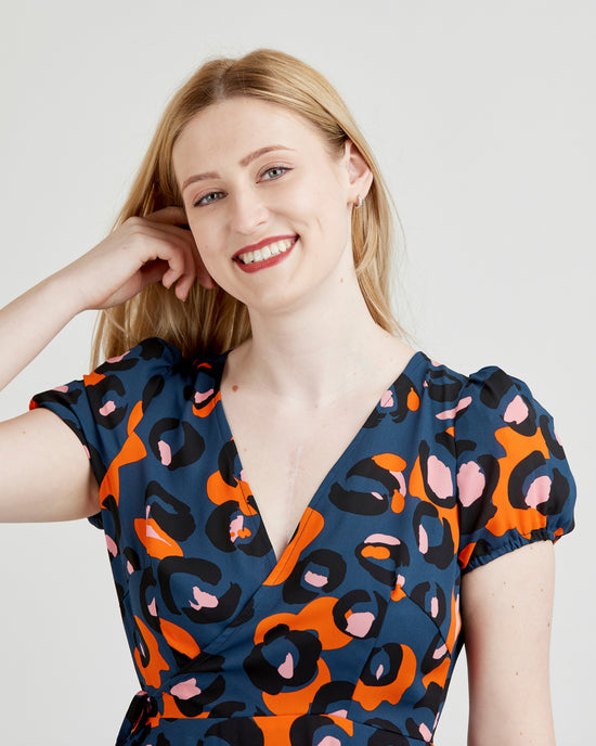 Woman wearing a blue dress with orange and pink abstract pattern on a plain background. This dress is the Roseclair Wrap Dress sewing pattern from Cashmerette.