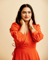 Woman wearing an orange dress with a beige background. This dress is the Roseclair Wrap Dress sewing pattern from Cashmerette.