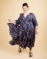 Woman wearing a navy dress with wavy pattern on a beige background. This dress is the Roseclair Wrap Dress sewing pattern from Cashmerette.