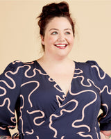 Woman wearing a navy dress with white pattern against a beige background. This dress is the Roseclair Wrap Dress sewing pattern from Cashmerette.