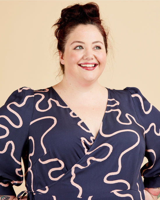 Woman wearing a navy dress with white pattern against a beige background. This dress is the Roseclair Wrap Dress sewing pattern from Cashmerette.