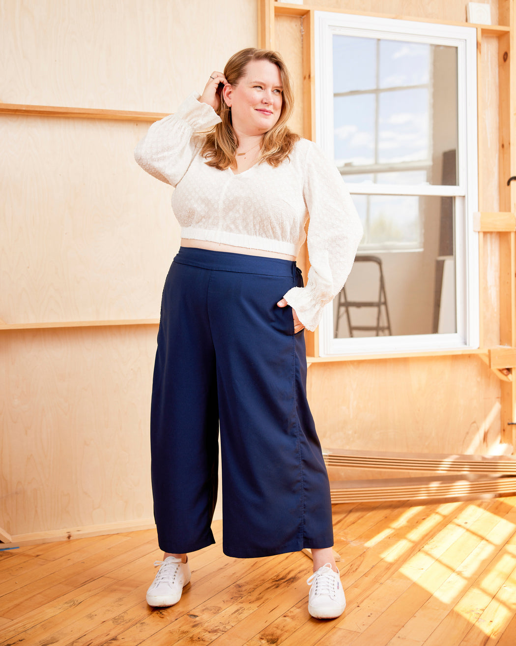 Woman wearing a white blouse and blue pants standing in a room with wooden floors and a window. This top is the Kineton Dress & Top sewing pattern from Cashmerette.