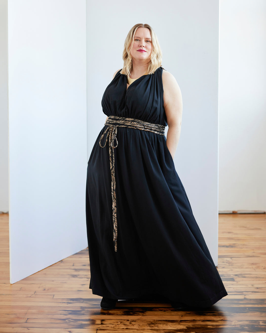 Woman wearing a black dress with a belt in a room with wooden flooring and white walls. This dress is the Rockwell Dress sewing pattern from Cashmerette.