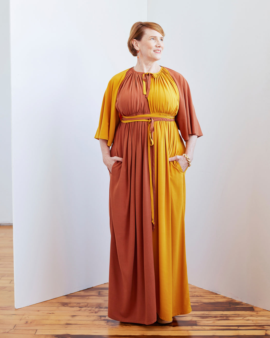 Woman wearing a two-tone dress with a yellow top and brown skirt against a white wall. This dress is the Rockwell Dress sewing pattern from Cashmerette.