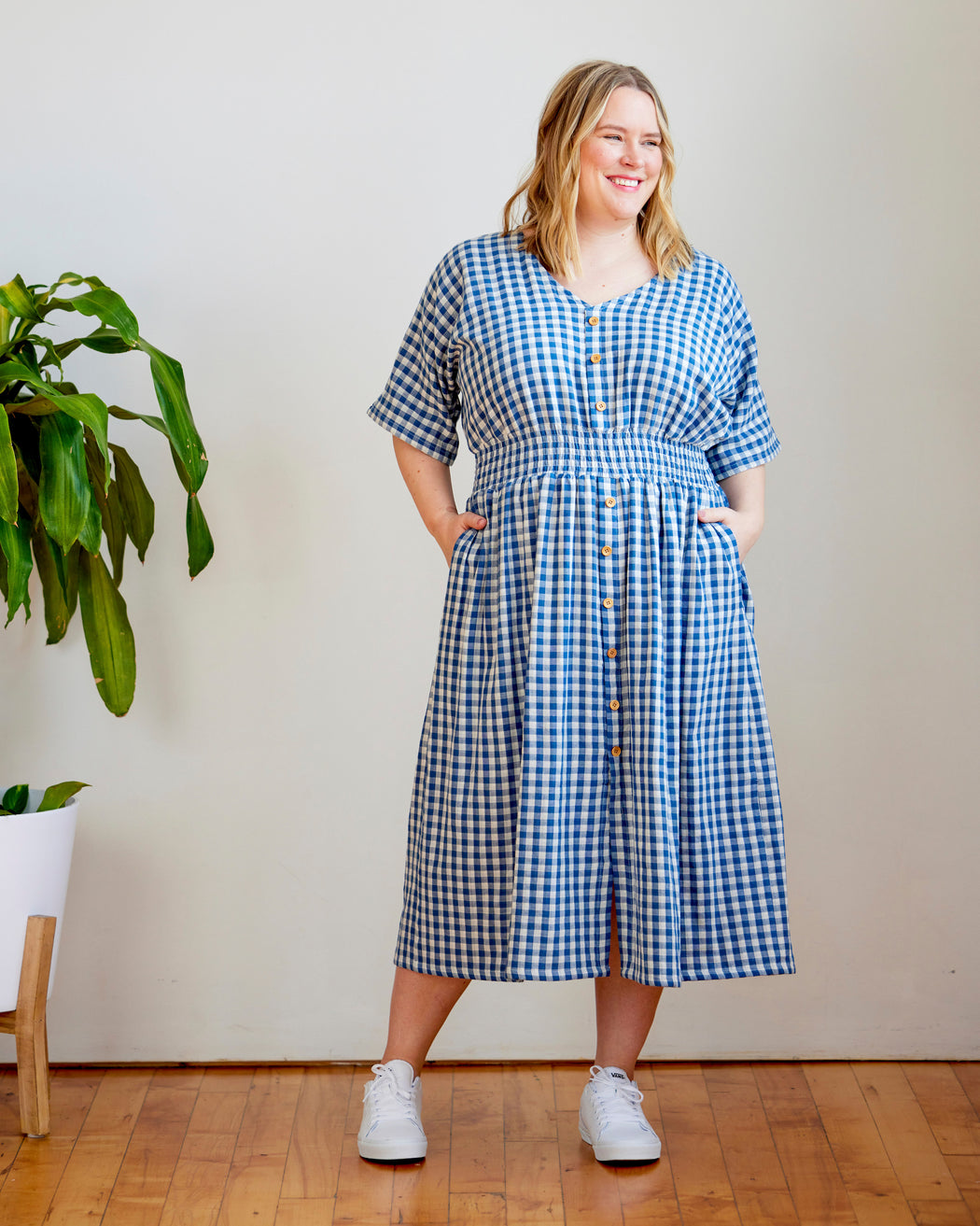 Woman wearing a blue checkered dress standing in a room with a plant and wooden floor. This dress is the Franconia Dress sewing pattern from Cashmerette.