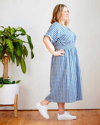 Woman wearing a blue and white checkered dress standing in a room with a plant and wooden floor. This dress is the Franconia Dress sewing pattern from Cashmerette.