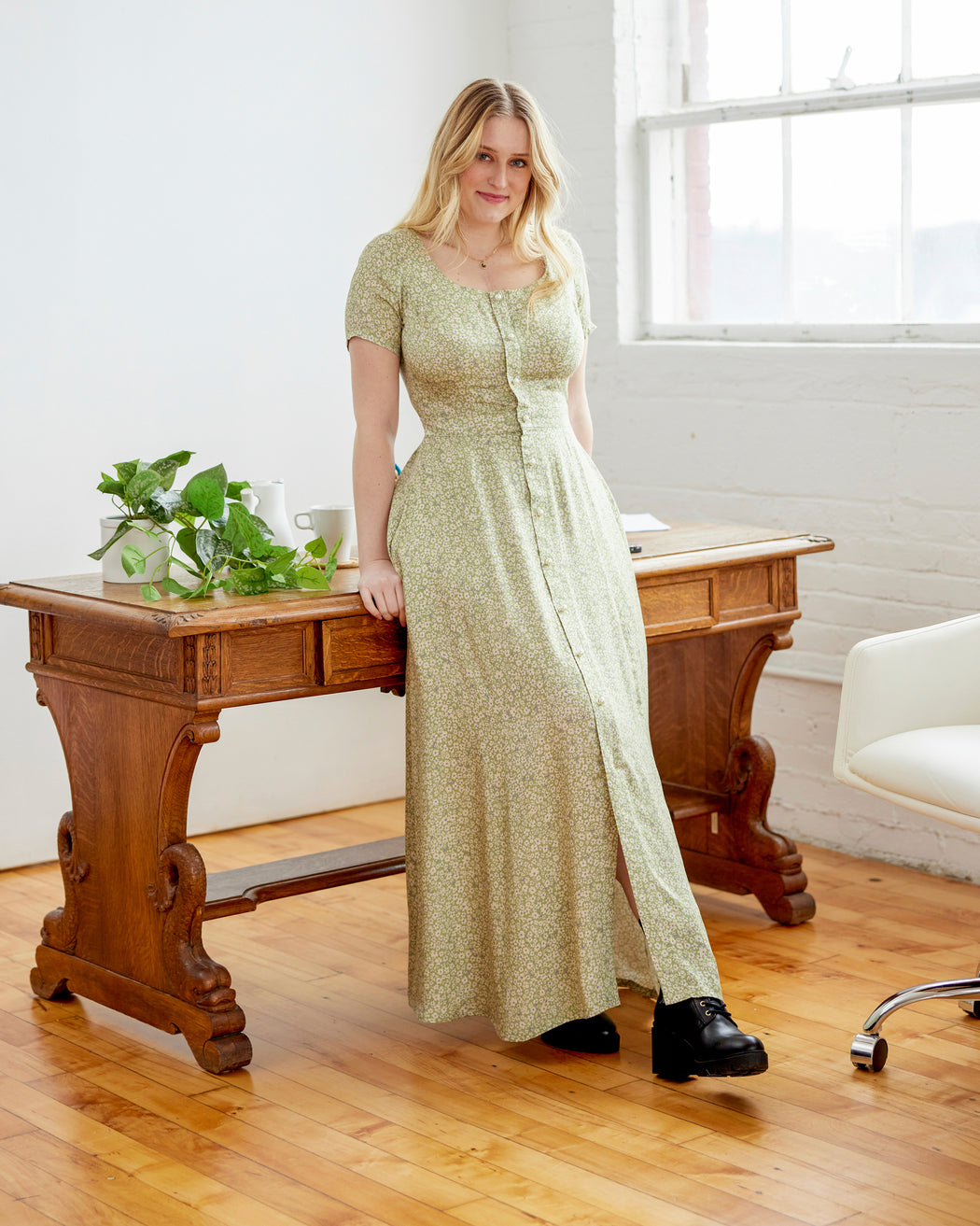 Woman in a long green dress standing in a room with wooden furniture and large windows. This dress is the Hampden Dress sewing pattern from Cashmerette.