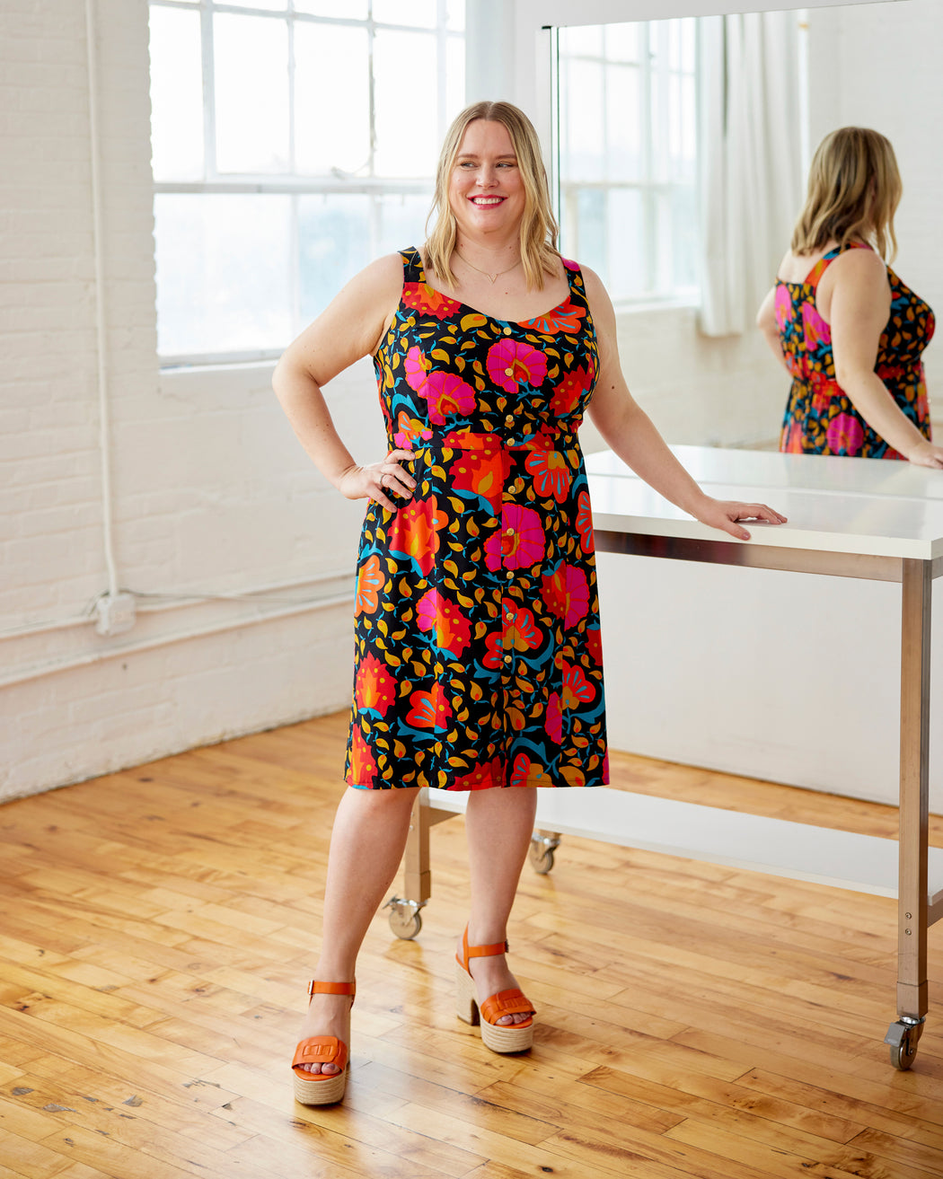 Woman wearing a colorful floral dress standing in a room with wooden floor and large windows. This dress is the Hampden Dress sewing pattern from Cashmerette.
