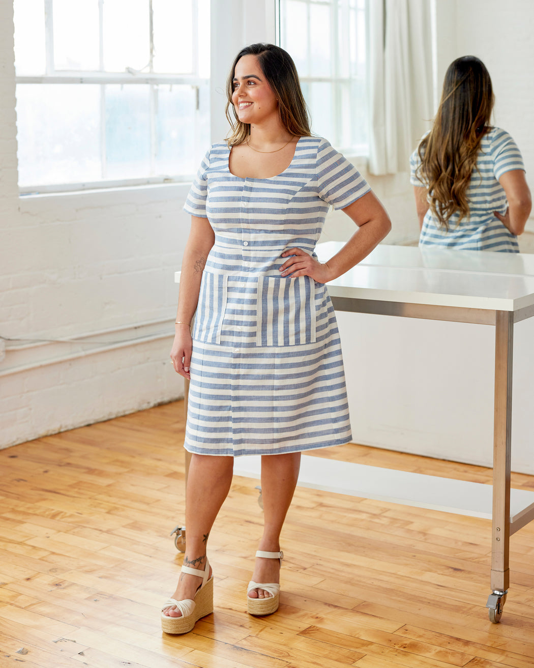 Woman wearing a blue and white striped dress standing in a bright room with large windows. This dress is the Hampden Dress sewing pattern from Cashmerette.