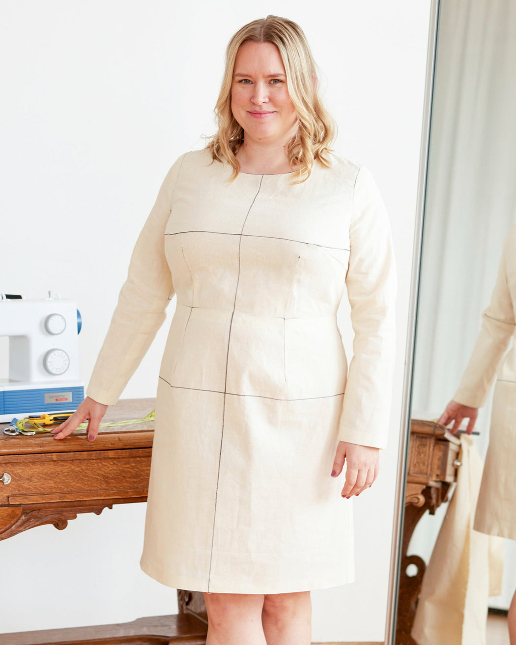 Woman wearing a beige dress standing next to a mirror in a room with a white wall and wooden furniture. This dress is the Wyman Dress Sloper sewing pattern from Cashmerette.
