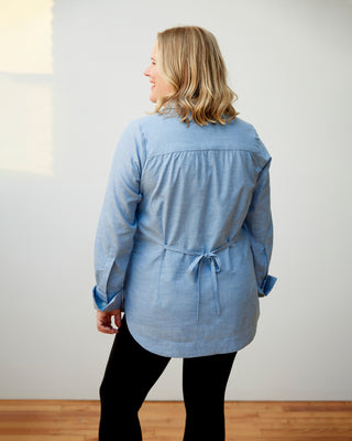 Woman wearing a light blue shirt with a tie waist against a white wall. This dress is the Winvale Dress & Tunic sewing pattern from Cashmerette.