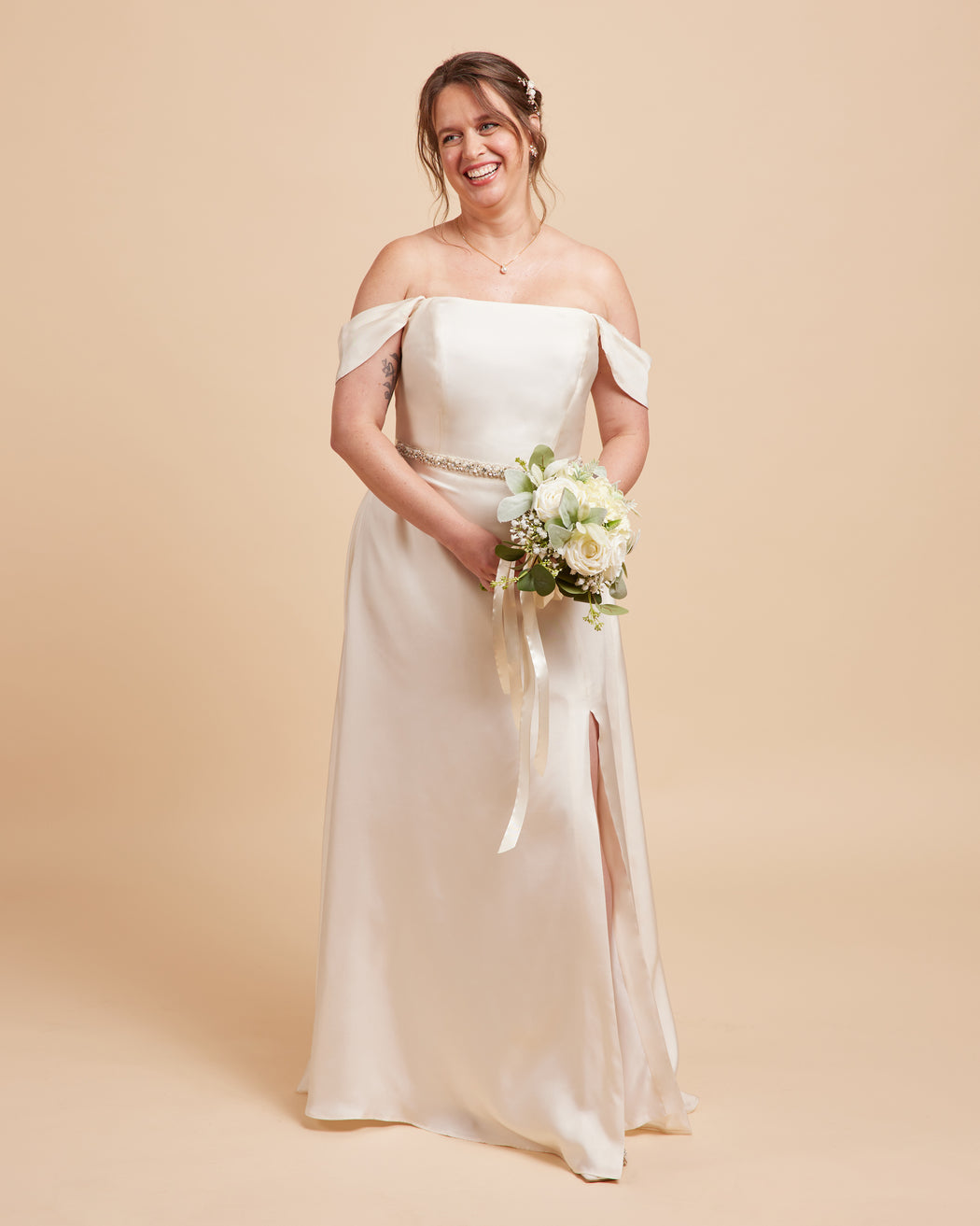 Woman in a white off-shoulder wedding dress holding a bouquet on a beige background. This dress is the Ellington Strapless Dress sewing pattern from Cashmerette.