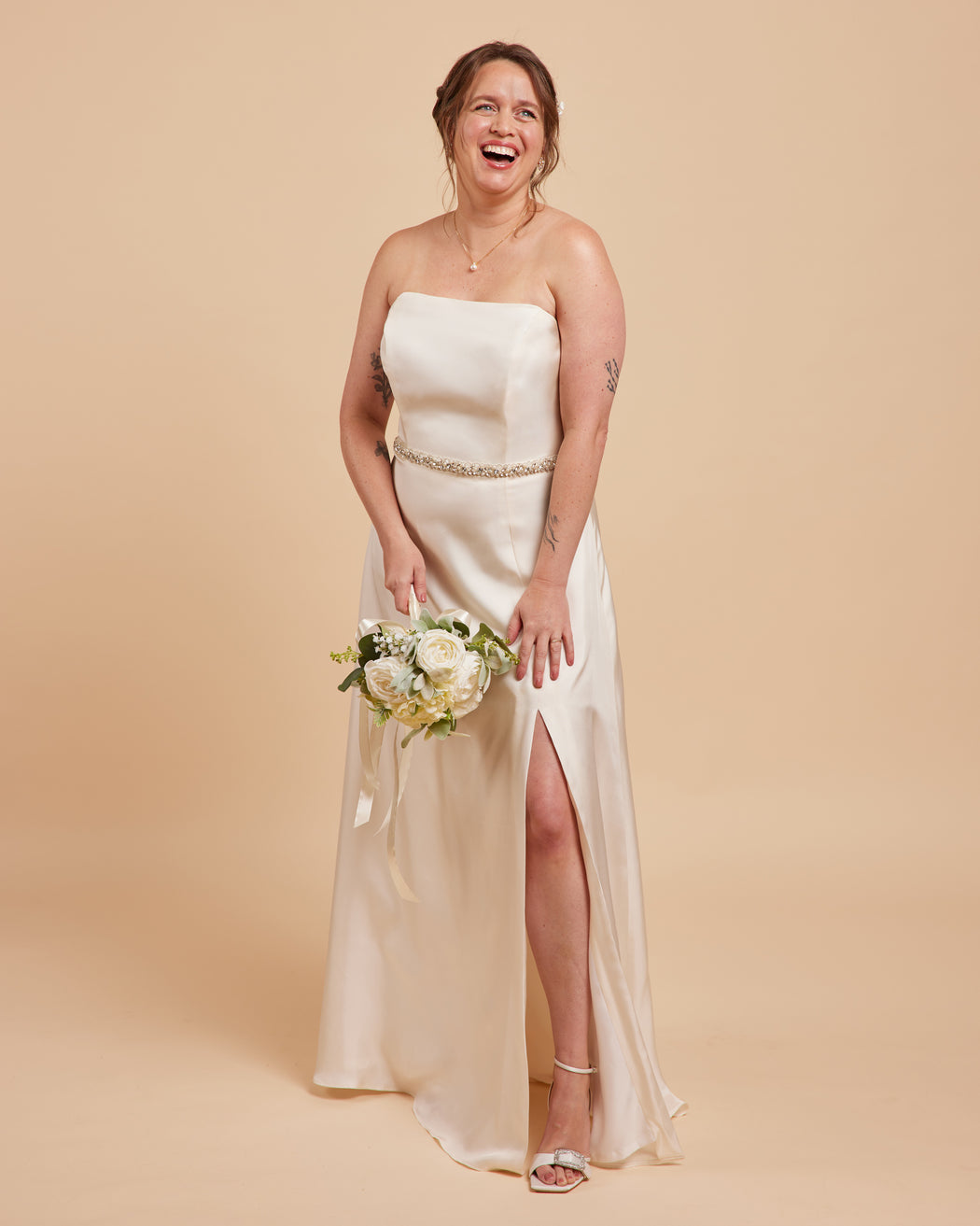 Woman in a strapless white wedding dress with a high slit, holding a bouquet on a beige background. This dress is the Ellington Strapless Dress sewing pattern from Cashmerette.