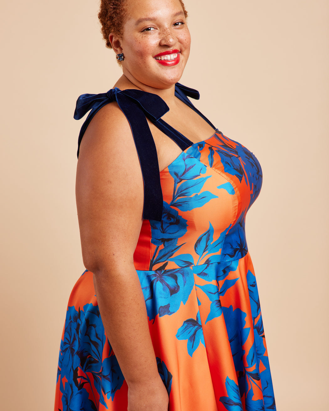 Woman wearing a colorful floral dress with a bow detail on a beige background. This dress is the Ellington Strapless Dress sewing pattern from Cashmerette.