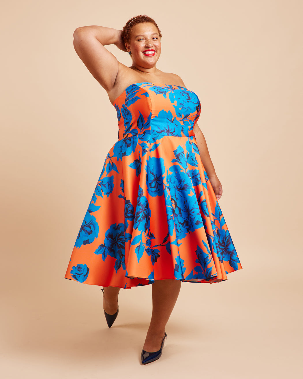 Woman wearing a strapless dress with orange and blue floral pattern on a beige background. This dress is the Ellington Strapless Dress sewing pattern from Cashmerette.