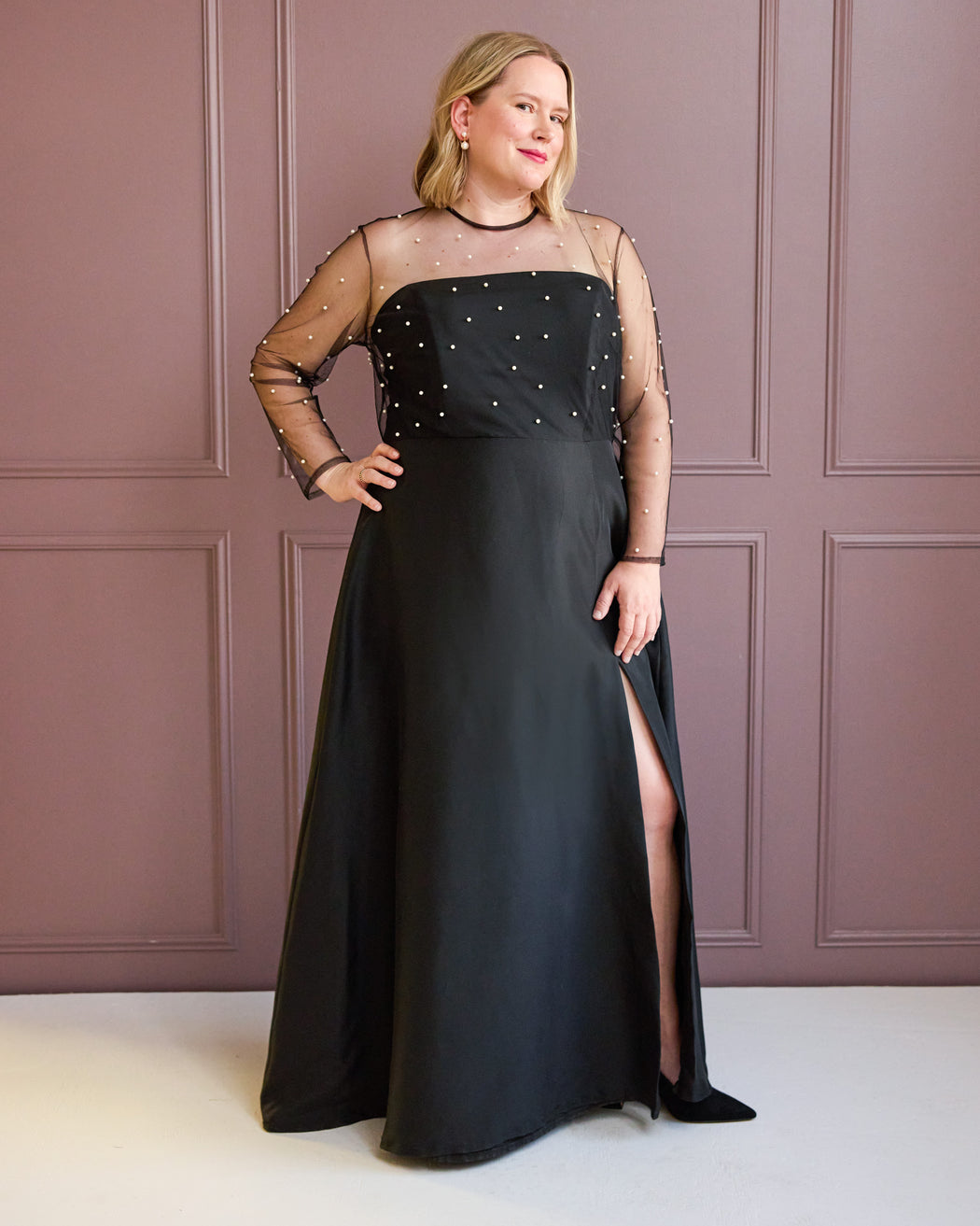 Woman wearing a black evening gown with sheer sleeves against a pink paneled wall. This is the Ellington Dress sewing pattern from Cashmerette.