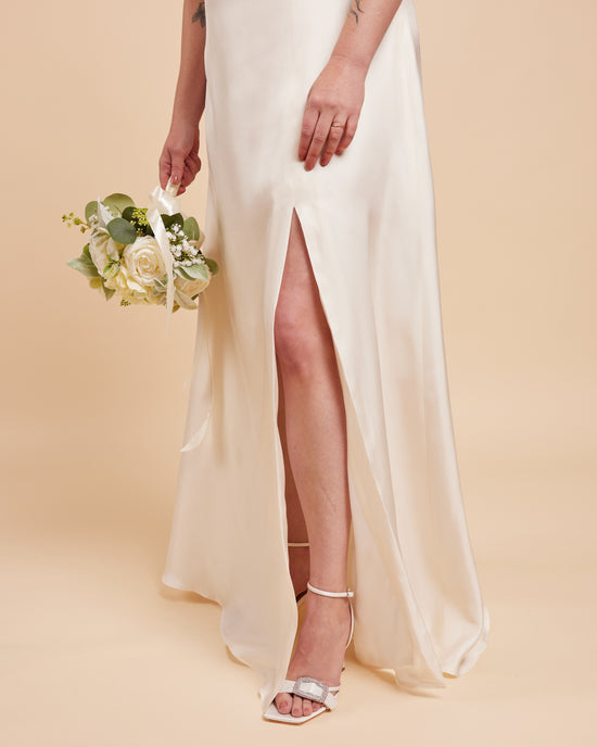 Woman in a white wedding dress with a high slit holding a bouquet on a beige background. This dress is the Ellington Strapless Dress sewing pattern from Cashmerette.