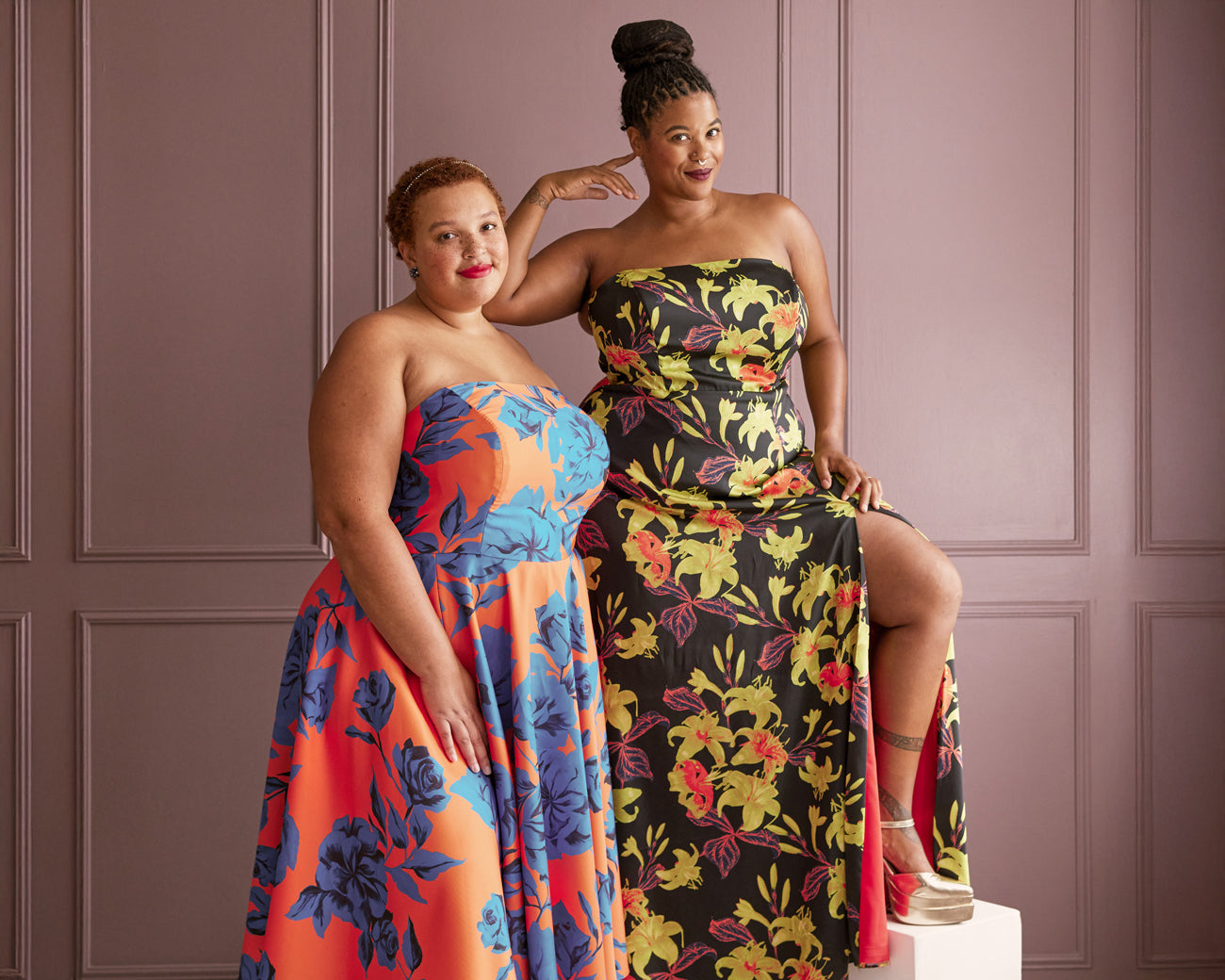 Two women in floral strapless dresses posing against a purple paneled wall.