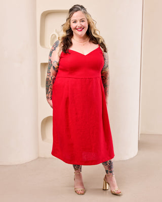 A woman modeling a bright red sleeveless midi dress with a sweetheart neckline and a flared skirt, standing against a minimalist beige architectural background. This is the Wayland Dress sewing pattern from Cashmerette.
