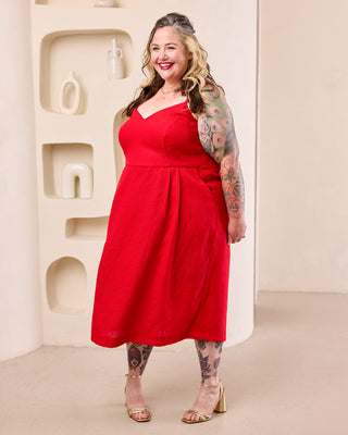 A woman modeling a bright red sleeveless midi dress with a V-neckline and gathered waist, standing in a minimalist beige interior setting. This is the Wayland Dress sewing pattern from Cashmerette.