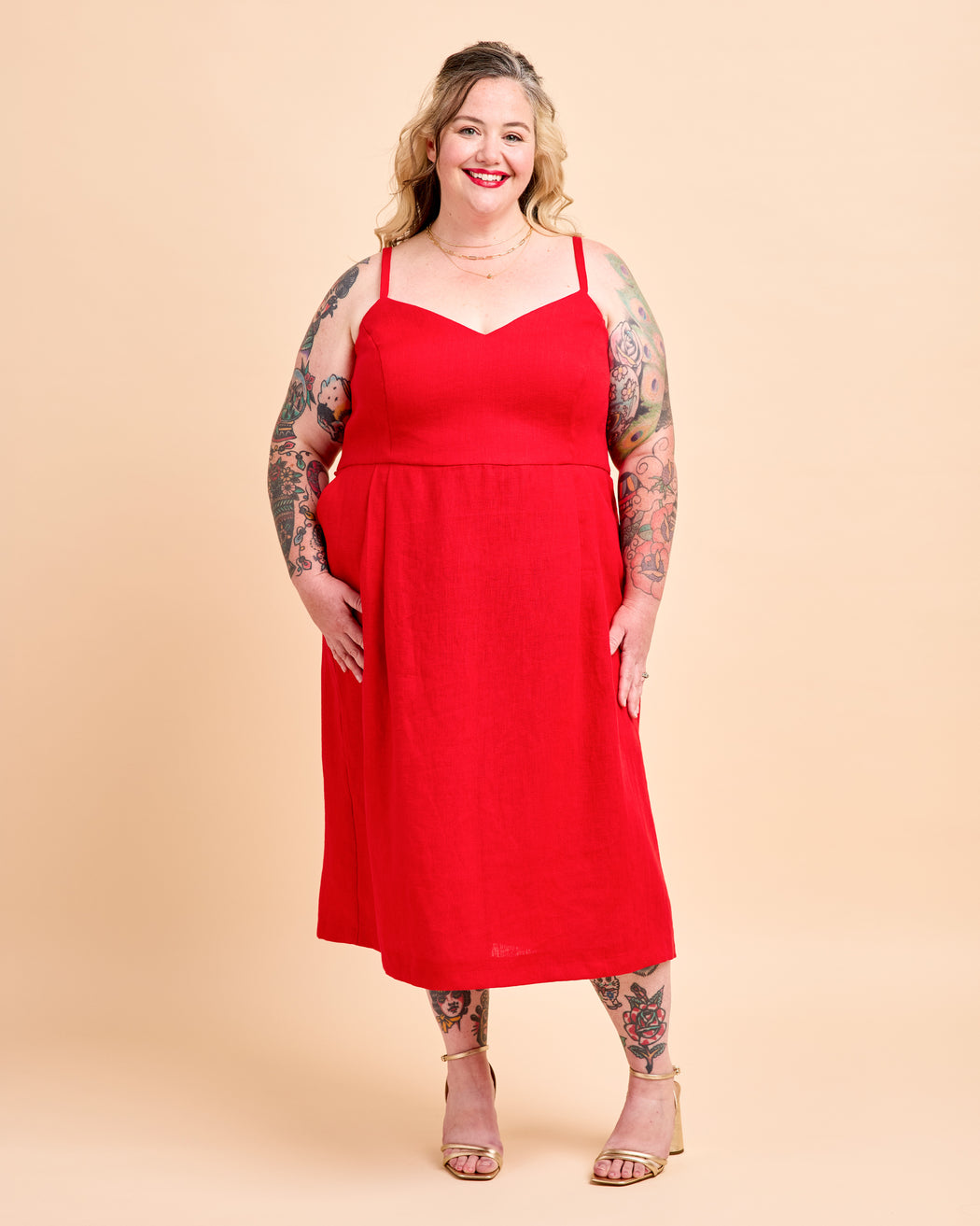 A plus-size woman with visible tattoos models a bright red midi dress with thin spaghetti straps and a V-neckline, standing against a solid beige studio background. This is the Wayland Dress sewing pattern from Cashmerette.