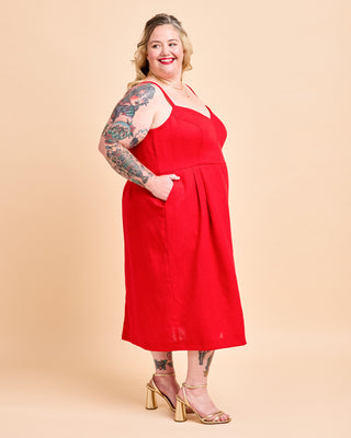 A plus-size model with tattoos is wearing a bright red sleeveless midi dress with thin straps and side pockets, paired with gold block-heel sandals, against a solid beige studio background. This is the Wayland Dress sewing pattern from Cashmerette.