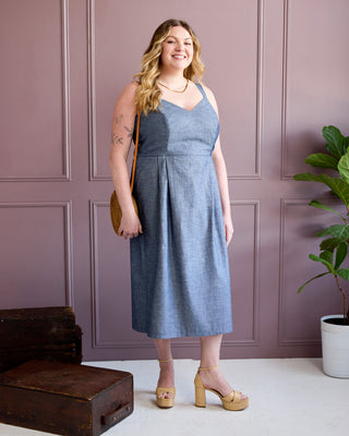A woman modeling a sleeveless, V-neck midi dress in a dusty blue chambray fabric, standing against a mauve paneled wall with a potted plant and a vintage wooden trunk. This is the Wayland Dress sewing pattern from Cashmerette.