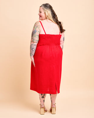 A woman modeling a bright red midi dress from the back, standing against a solid light beige studio background. This is the Wayland Dress sewing pattern from Cashmerette.
